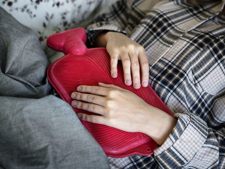  Das Bild zeigt eine Person, die entspannt auf einer Couch oder einem Bett liegt, mit einer rot gefärbten Wärmflasche auf ihrem Bauch. Die Person trägt eine karierte Jacke und liegt unter einer grauen Decke. Ihre Hände liegen sanft auf der Wärmflasche, was darauf hindeuten könnte, dass sie Wärme und Komfort sucht, möglicherweise um Schmerzen oder Unbehagen zu lindern. Das Muster der Jacke und die Textur der Wärmflasche sind deutlich sichtbar und erzeugen einen gemütlichen und behaglichen Eindruck.