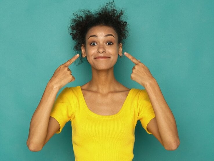  Ein Bild einer Frau in einem gelben T-Shirt, die lächelnd ihre Zeigefinger an beiden Seiten ihres Kopfes positioniert, um auf ihre Ohren zu zeigen. Sie steht vor einem einfarbigen türkisfarbenen Hintergrund, was einen starken Kontrast zu ihrem gelben Oberteil bildet. Ihr Ausdruck ist heiter und spielerisch.
