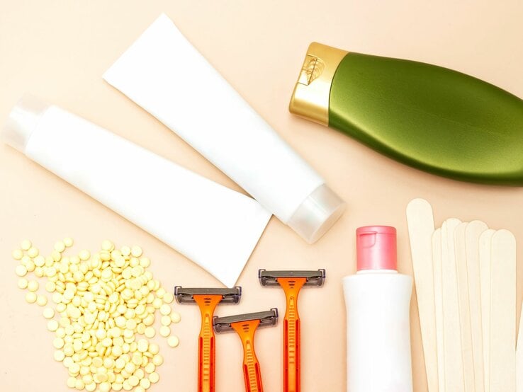Eine Reihe von Hygieneprodukten auf einem Tisch, darunter gelbes Wachs in einem Behälter, ein Holzstäbchen, ein Rasierer, sowie Flaschen mit Intimlotion und -shampoo, alle arrangiert für die persönliche Pflege.