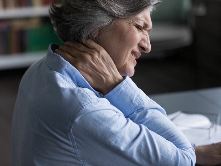 Eine ältere Frau fasst sich mit der Hand an den Hals und verzieht vor schmerz das Gesicht. Sie hat einseitige Halsschmerzen.