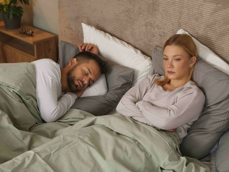 Verärgerte Frau denkt neben ihrem schlafenden Partner über Beziehungsprobleme und die Gleichgültigkeit des Partners nach.