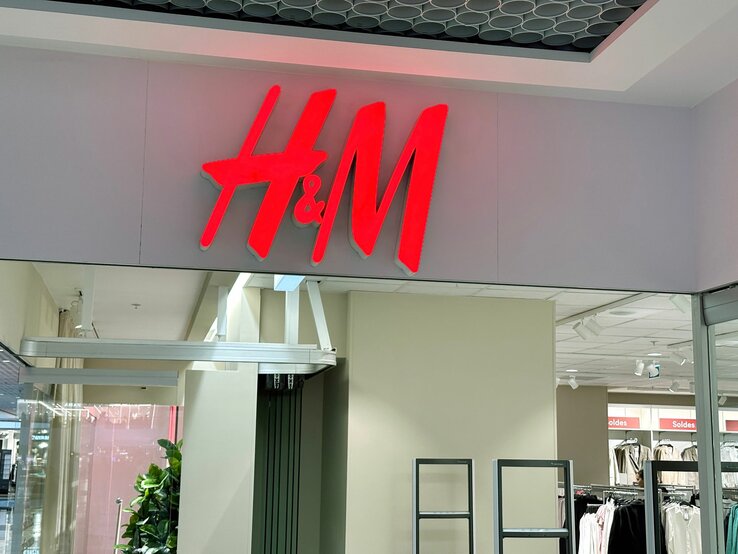 Leuchtschild des H&M-Logos in Rot über dem Eingang einer Filiale mit sichtbarem Innenraum und Kleidungsständern.