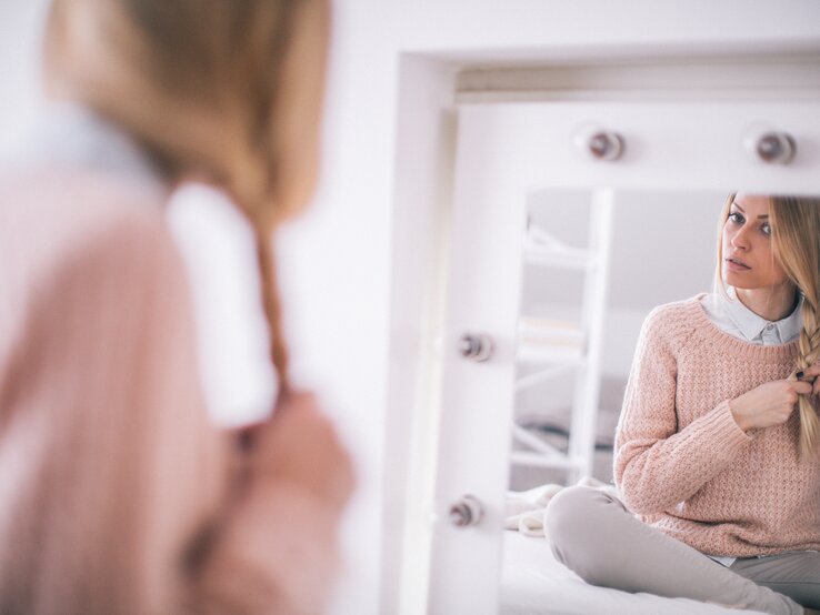 Frau, die vor einem Spiegel sitzt und sich selbst dabei betrachtet, wie sie ihr blondes Haar zu einem Zopf flicht. Sie trägt einen gemütlichen rosafarbenen Pullover und darunter ein hellblaues Hemd