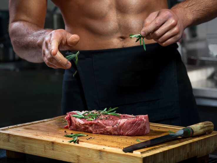 Cedrik Lorenzen: Ein durchtrainierter Mann mit freiem Oberkörper bereitet ein Stück Steak in der Küche zu.