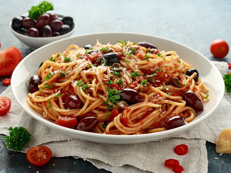 Ein weißer Pastateller mit den Spaghetti alla Puttanesca auf grauem Untergrund mit weißem Leinentuch. Daneben diverse Pastazutaten wie Parmesan, Tomaten, Oliven, Chili und Petersilie.