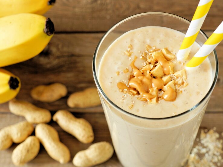 Erdnuss-Bananen-Shake mit Haferflocken im Glas mit Strohhalm und Toppings auf Holztisch. Daneben Erdnüsse und Bananen. Draufsicht.