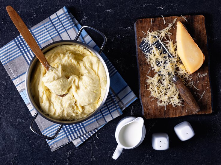 Käse-Kartoffelpüree in Topf auf dunkelblauem Untergrund. Daneben geriebener Käse.