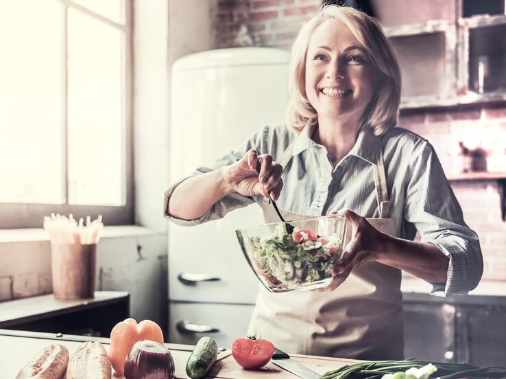 Eine lächelnde Frau in Küchenschürze mischt einen frischen Salat in einer Schüssel, umgeben von verschiedenen Gemüsesorten auf einer Küchenarbeitsplatte.
