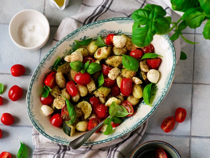 Gnocchi-Salat Caprese mit Tomaten, Zucchini und Mozzarella in kitschiger Schale auf weißen Fliesen. Daneben Basilikum, Salz, kirschtomaten und Olivenöl.