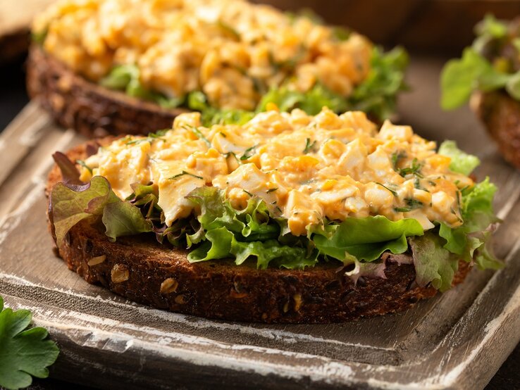 Zwei Brote mit dem Eiersalat auf Salatblättern. Alles auf einem dunkelbraunen Schneidebrett und rustikaler Tischplatte seitlich fotografiert.