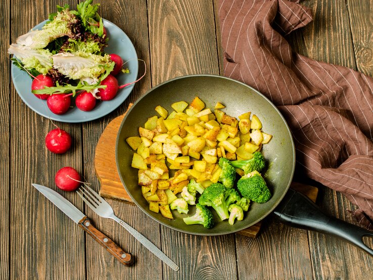 Draufsicht: Untergrund aus Holz, eine Pfanne  Bratkartoffeln mit Brokkoli sowie ein Salat mit Radieschen und Lollo Rosso-Salat. 