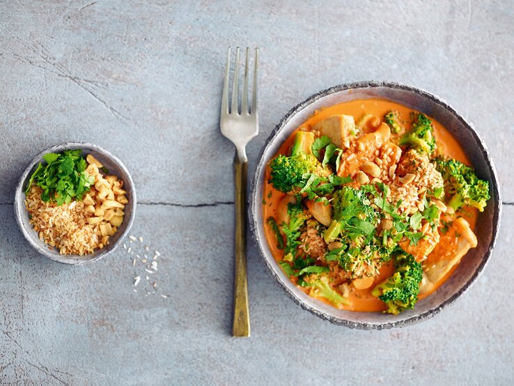 Ein Teller Hähnchen-Brokkoli-Curry in der Draufsicht, daneben eine Gabel und eine Schale mit Erdnüssen und Kräutern.