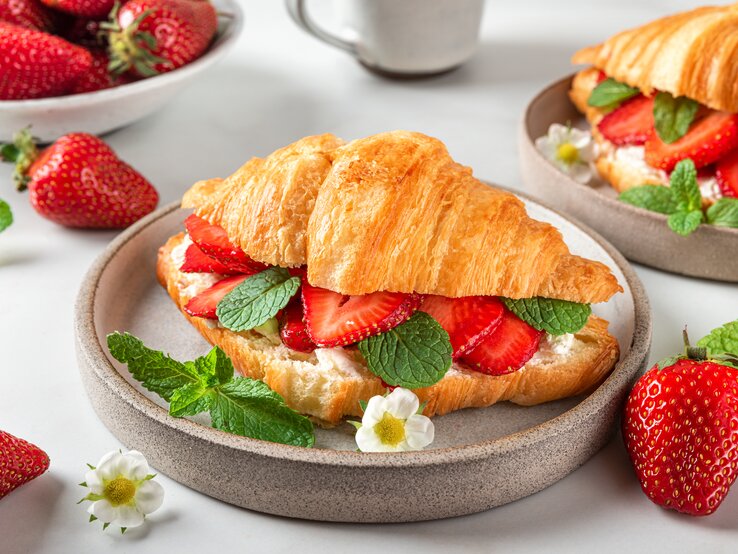 Frontalsicht: Ein Erdbeer-Croissant mit frischer Minze liegt in einer runden Tonschüssel, die auf einem weißen Untergrund liegt. Drum herum liegen viele frische Erdbeeren und weiße Erdbeerblüten.