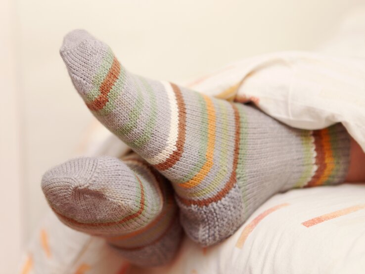 Eine Person liegt entspannt im Bett mit den Füßen hoch, die in gestreiften Socken in sanften Grau-, Grün-, Orange- und Weißtönen gehüllt sind. Der Fokus liegt auf den gemütlichen Socken, die gegen die weiche Bettwäsche drücken.