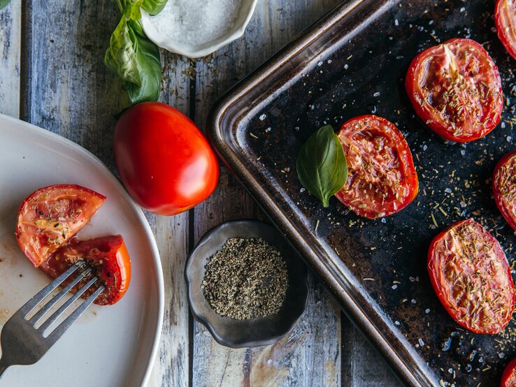Au der Vogelperspektive: Auf einem Ofenblech liegen halbierte Tomaten, die mit Kräutern betreut sind. Daneben steht ein weißer Teller, auf dem ebenfalls eine halbierte Tomate liegt.