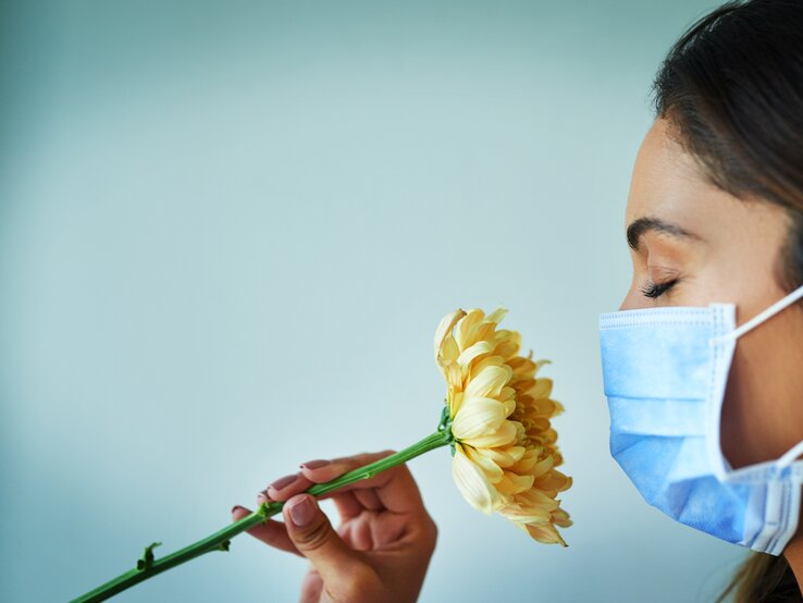 Jemand riecht an einer Blume, trägt dabei aber eine Atemschutzmaske. | © Getty Images/PeopleImages