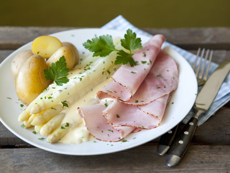 Spargel-Sauce Hollandaise Ein weißer Teller mit Spargel, Kartoffeln, Schinken und Sauce hollandaise auf einem rustikalen Holztisch.