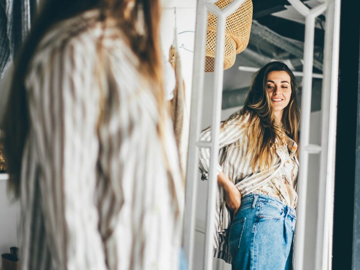 Junge Frau, die sich in einem großen, stehenden Spiegel betrachtet. Sie trägt eine lässige Jeans und eine gestreifte Bluse.  | © Adobe Stock / Mariia Korneeva