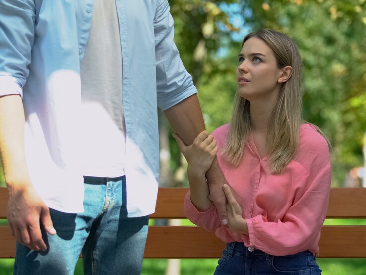 Mann lässt verärgerte Freundin allein auf Bank im Park zurück. Frau hält den Arm des Mannes fest.