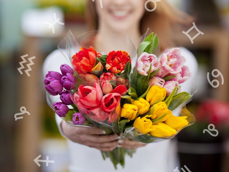  Eine junge Frau, die unscharf im Hintergrund lächelt, hält ein farbenfrohes Bouquet von Tulpen in den Händen. Die Blumen, in leuchtenden Tönen von Lila, Rot, Rosa und Gelb, sind frisch und in Klarsichtfolie eingewickelt, wobei der Fokus auf den Tulpen im Vordergrund liegt. Umgeben ist sie von einem Kreis mit den astrologischen Symbolen der Tierkreiszeichen.