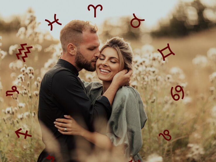 Ein glückliches Paar umarmt sich in einem Feld mit weißen Blüten. Der Mann küsst die Frau sanft an der Schläfe. Um sie herum sind astrologische Symbole wie Wassermann, Widder und andere verteilt.