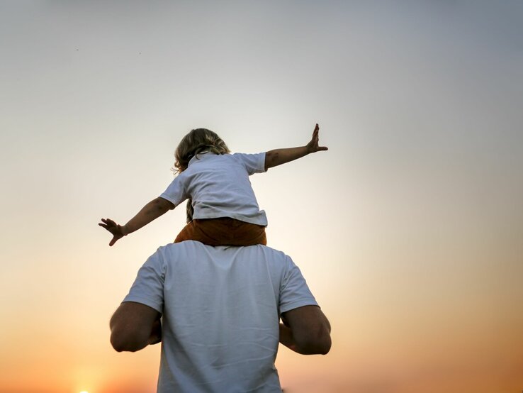 Kind im Flugzeugspiel auf Schultern eines Mannes gegen den Himmel im Sonnenuntergang, strahlt Freiheit aus.