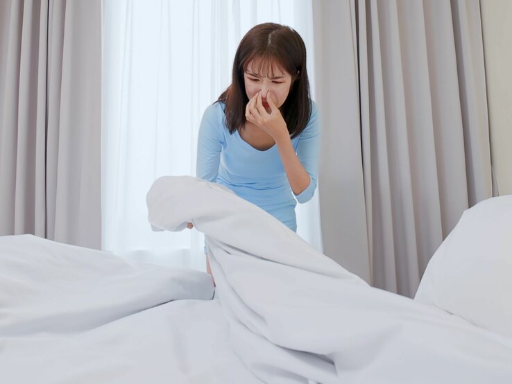 Eine Frau hält sich die Nase zu, während Sie Bettwäsche auf dem Bett hochhebt.