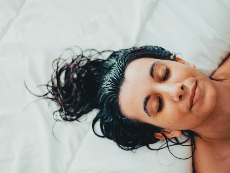 Junge Frau, die entspannt mit geschlossenen Augen und nassen Haaren auf einem weißen Bettlaken liegt.