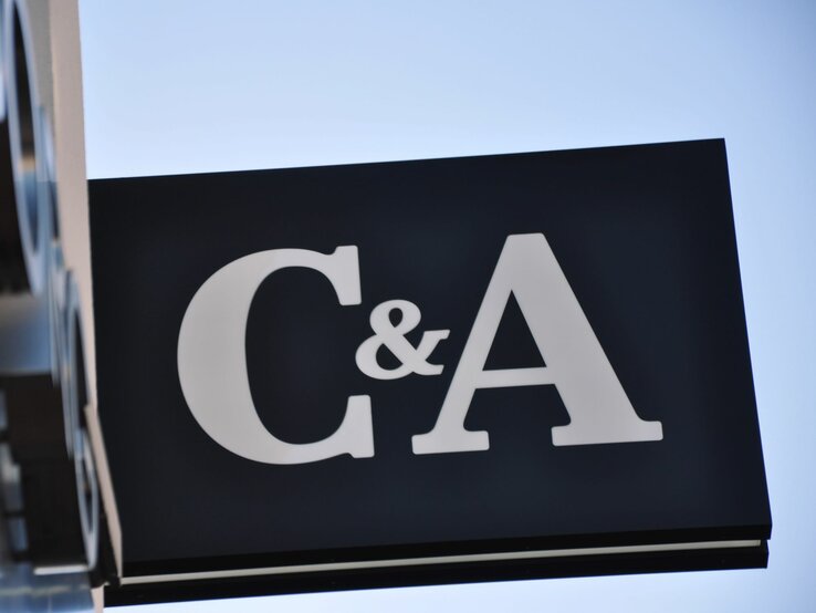  Ein großes, schlichtes Schild mit den Buchstaben "C&A" in weißer Farbe auf einem schwarzen Hintergrund. Das Schild ist schräg fotografiert gegen einen klaren blauen Himmel. Die Sonne scheint von der Seite und beleuchtet das Schild und seine Kanten.