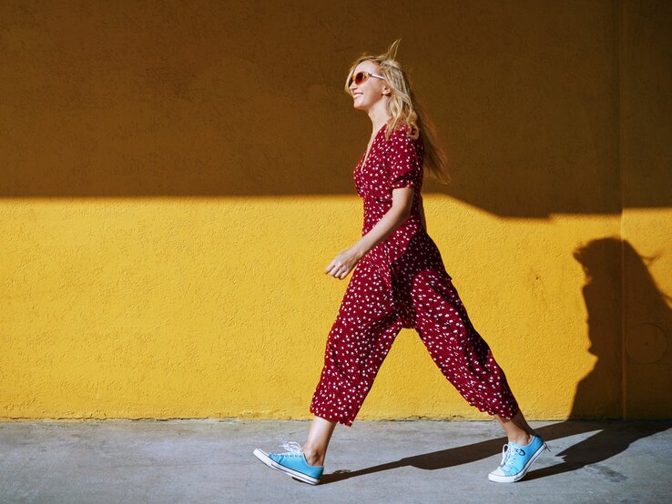 Stilvolle blonde Dame in rot-weiß gepunktetem Jumpsuit und blauen Sneakern läuft lächelnd an einer gelben Wand entlang.