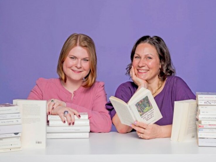 Zwei Frauen sitzen vor einem lila Hintergrund und haben einen Stapel Bücher vor sich.