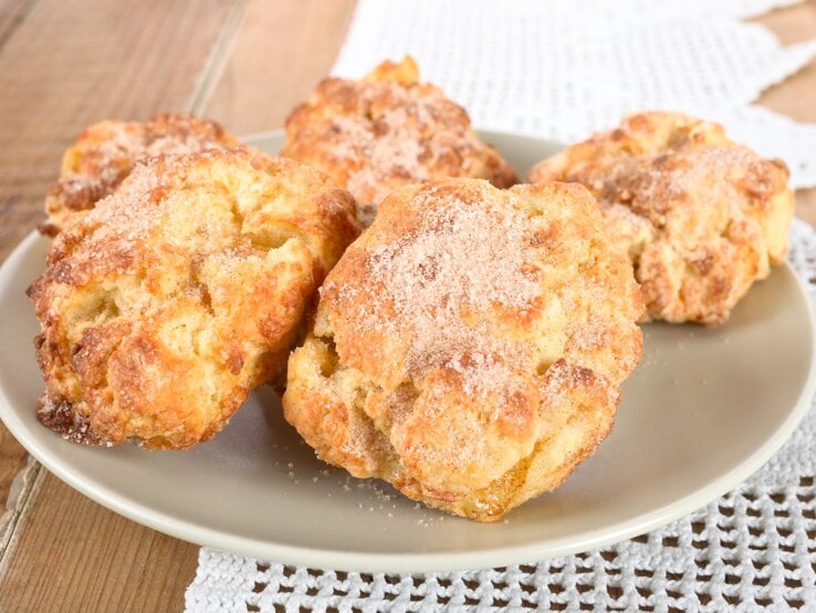 Kleines Gebäck ist immer schnell vernascht. Unsere saftigen Apfel-Quark-Taler sind aber genauso schnell gebacken und daher immer für den kleinen Hunger zur Stelle. Gebackene Apfel-Quark-Taler auf einem Teller auf Holzunterlage, dahinter Tischdeckchen