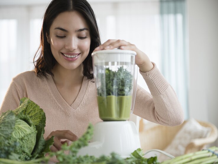 Eine lächelnde junge Frau bereitet in ihrer Küche einen grünen Smoothie zu. Sie legt ihre Hand auf den Deckel eines Mixers, der mit grünen Blättern gefüllt ist, vermutlich Kale oder Spinat. Vor ihr auf dem Tisch liegen frische grüne Gemüsesorten. Das Bild vermittelt ein Gefühl von Gesundheit und Wohlbefinden durch die Zubereitung von frischen, nahrhaften Lebensmitteln. Die Küche ist hell und einladend mit einem Fenster im Hintergrund.