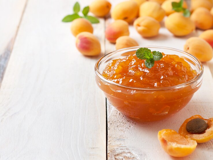 Auf weißen Holzbrettern steht eine Glasschale mit Marillen-Konfitüre. Drumherum liegen einzelne Marillen.