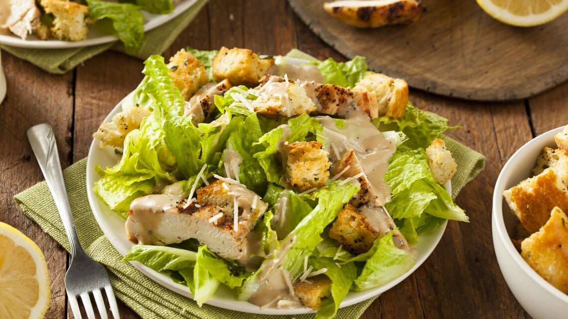 Caesar Salad mit gegrillter Putenbrust und Salatblättern / runder Teller / Gabel / Stoffserviette / angeschnittene Zitrone / Draufsicht, schräg
