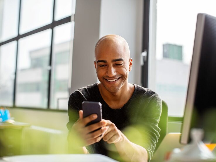 Junger Mann mit Glatze lacht am Schreibtisch beim Blick in sein Handy