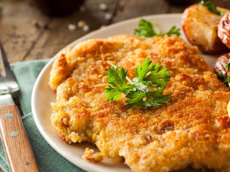 Ein Schnitzel auf einem Teller mit Ofenkartoffeln, Petersilie und Zitrone. Alles auf einem rustikalen Holztisch mit dunkelgrüner Tischdecke und Gabel. Sehr nah von der Seite fotografiert.