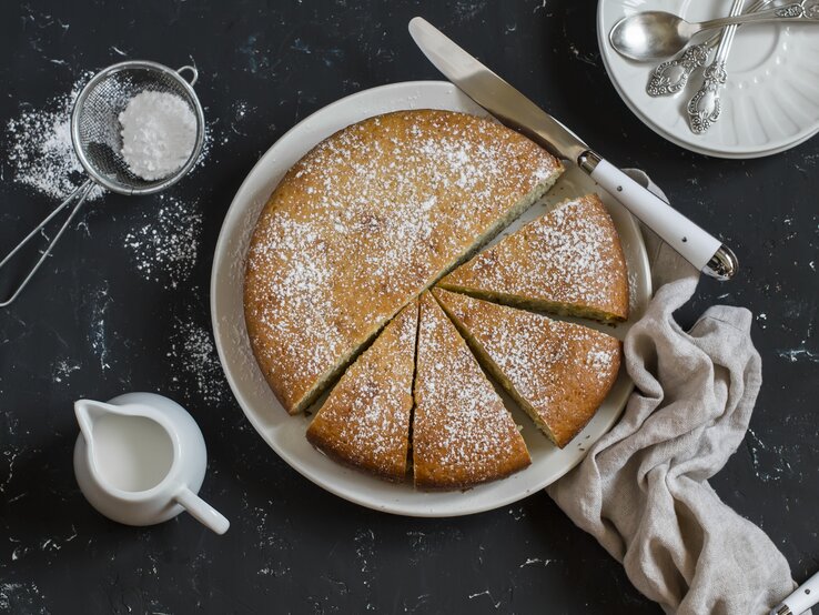 Runder, heller Kuchen bestäubt mit Puderzucker auf weißem Teller. Dieser steht auf einer dunklen Arbeitsfläche. 