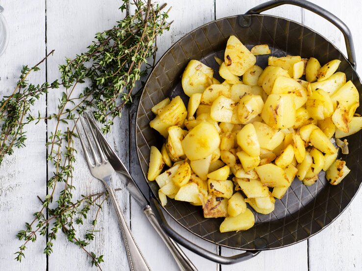 Gusseiserne Pfanne gefüllt mit kross gebratenen Kartoffeln auf weißem Holztisch. 