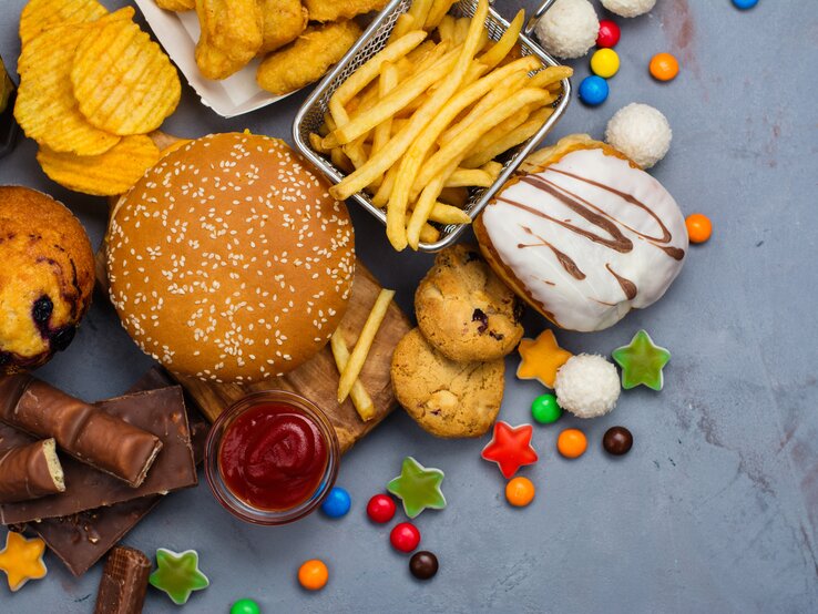 Pommes, Burger, Gummitierchen und Kuchen liegen auf einem Tisch. Diese Lebensmittel fördern eine Fettleber. 