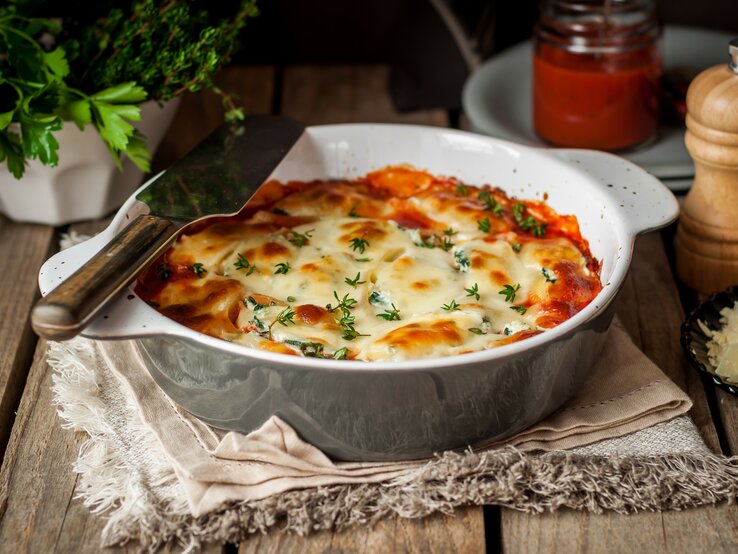 Garnelenauflauf mit Tagliatelle und 3-Käse-Béchamel in Auflaufform auf Tuch auf Holztisch. Daneben Kräuter, geriebener Käse, ein Glas mit Soße und Pfeffermühle. Frontalansicht.