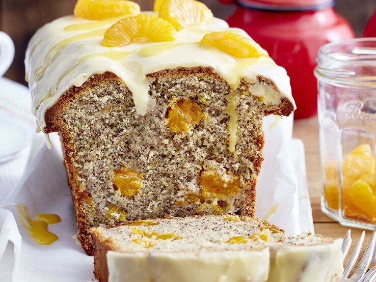 Auf einem Tisch steht ein angeschnittener Mohn-Eierlikör-Kastenkuchen. Er ist mit Mandarinen bedeckt. 