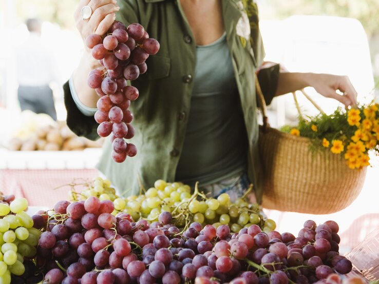 Das Bild zeigt eine Frau, die eine Traube roter Trauben über einem Marktstand hält, der mit verschiedenen Traubensorten gefüllt ist. Die Trauben erscheinen frisch und saftig, mit einem Farbspektrum von hellgrün bis dunkellila. Die Frau trägt ein grünes Oberteil und hat einen geflochtenen Korb über dem Arm, der mit Blumen geschmückt ist, was darauf hindeutet, dass sie frische Produkte und Blumen einkauft. Der Fokus liegt auf den Trauben und den Händen der Person, während der Hintergrund mit anderen Marktaktivitäten unscharf bleibt, was eine lebendige Marktatmosphäre vermittelt. 