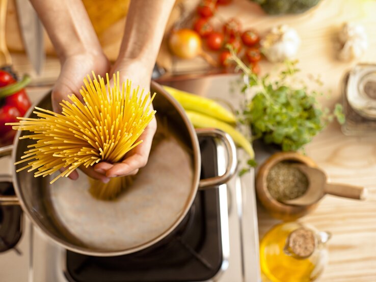 Spaghetti werden in einen Topf voll Wasser gegeben. Um den Herd herum liegen verschiedene, typisch italienische Zutaten. 