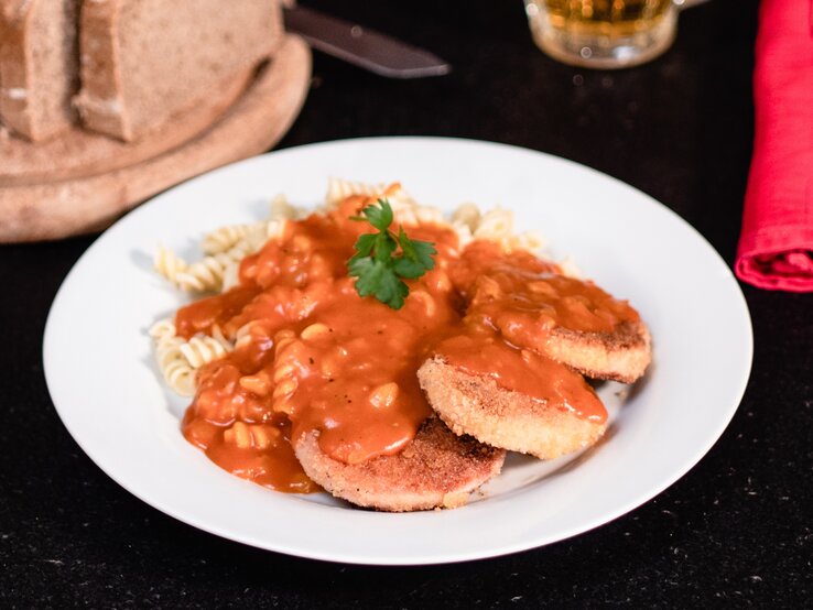 DDR Jägerschnitzel – panierte Jagdwurstscheiben mit Spirelli-Nudeln Tomatensoße. Ostdeutsches Gericht auf weißem Teller. Daneben angeschnittenes Brot und Besteck. 