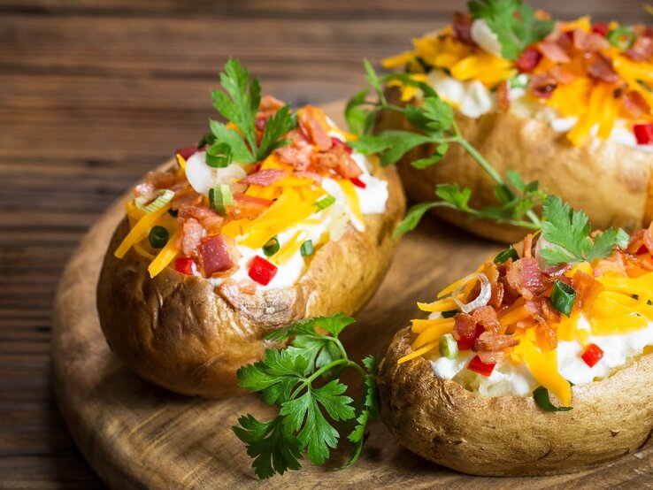 Drei Ofenkartoffeln auf einem Holzbrett. Die Kartoffeln sind gefüllt mit Kräuterquark und mit Petersilie, Käse und Speck bestreut.