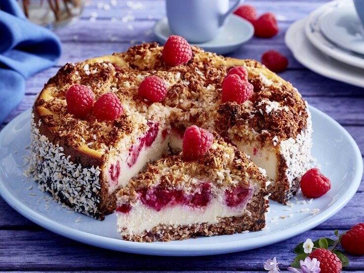 Auf einem Teller liegt ein Schmandkuchen mit Himbeeren. Er ist mit Himbeeren und Kokosraspeln dekoriert.