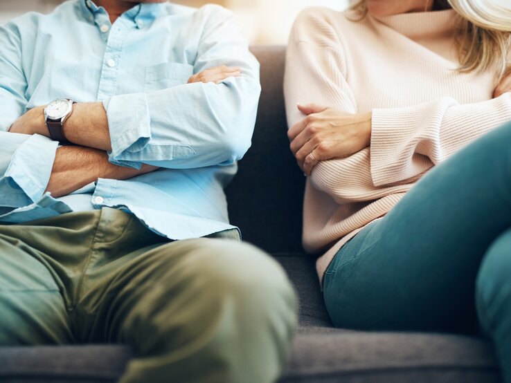 Mann und Frau sitzen nebeneinander mit verschränkten Armen auf einer Couch. Man sieht hauptsächlich den Körper, die Köpfe sind abgeschnitten.
