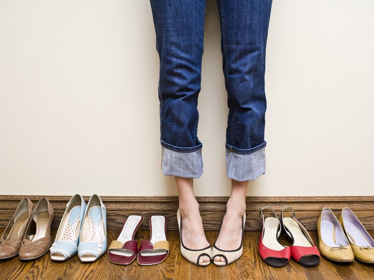 Viele Schuhe in einer Reihe aufgestellt, dazwischen die Beine und Füße einer Frau. 
