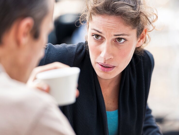 Frau schaut im Gespräch im Freien an einem Tisch im ungläubig auf Mann, er hält eine Kaffeetasse